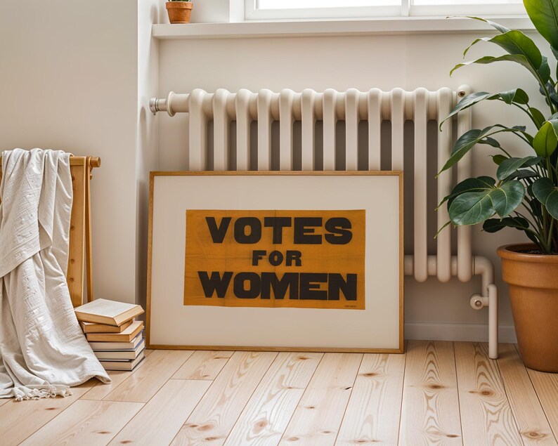 gs print shoppe, gsprintshoppe.com, vintage-style suffrage poster art print featuring bold black text 'VOTES FOR WOMEN' on an orange-yellow banner, framed in minimalist wood and displayed in bright modern interiors