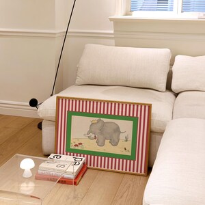 May include: A framed illustration of a gray elephant wearing a crown, set against a green and red striped background. The artwork is displayed on a light-colored sofa, with a modern black floor lamp and a clear acrylic side table.