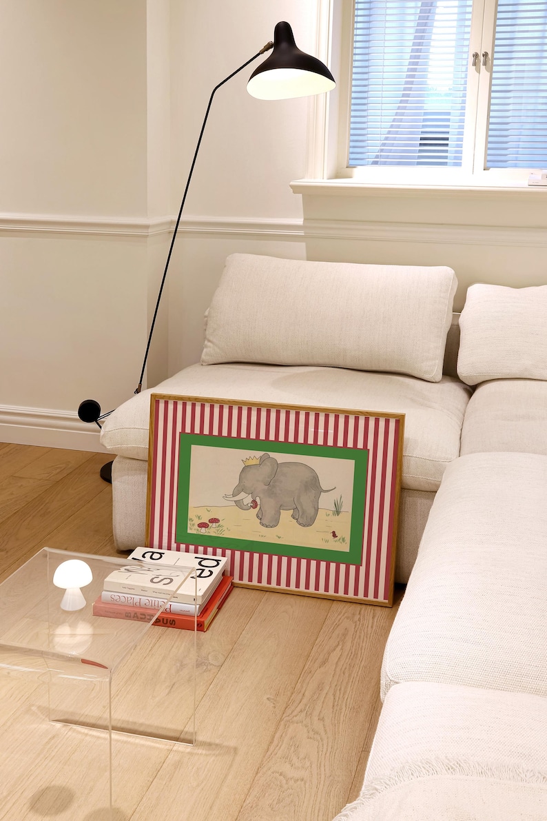 May include: A framed illustration of a gray elephant wearing a crown, set against a green and red striped background. The artwork is displayed on a light-colored sofa, with a modern black floor lamp and a clear acrylic side table.