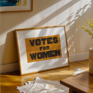 gs print shoppe, gsprintshoppe.com, vintage-style suffrage poster art print featuring bold black text 'VOTES FOR WOMEN' on an orange-yellow banner, framed in minimalist wood and displayed in bright modern interiors