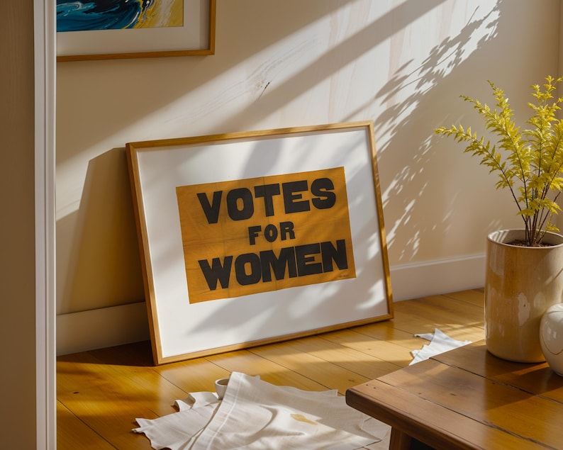 gs print shoppe, gsprintshoppe.com, vintage-style suffrage poster art print featuring bold black text 'VOTES FOR WOMEN' on an orange-yellow banner, framed in minimalist wood and displayed in bright modern interiors