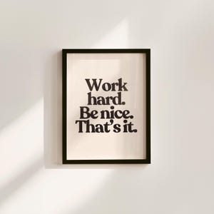 May include: A framed print with the text "Work hard. Be nice. That's it." in a bold, black font. The print has a cream-colored background and is encased in a black frame, suitable for home or office decor.