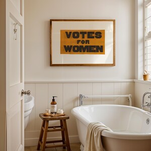 gs print shoppe, gsprintshoppe.com, vintage-style suffrage poster art print featuring bold black text 'VOTES FOR WOMEN' on an orange-yellow banner, framed in minimalist wood and displayed in bright modern interiors