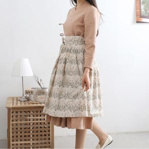 Puede incluir: Una falda beige y marr&oacute;n con estampado floral. La falda es plisada y tiene una cintura ancha. La falda se lleva con una blusa marr&oacute;n claro.