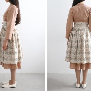 Puede incluir: Una blusa de manga larga de color marr&oacute;n claro con cuello alto y una falda blanca con estampado floral. La falda tiene una falda interior marr&oacute;n.