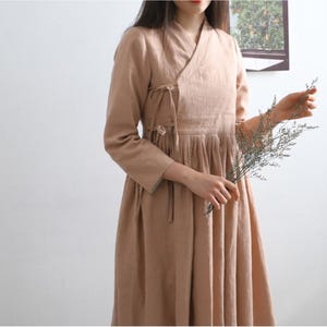 Puede incluir: Un vestido largo de lino de color marr&oacute;n claro con cuello en V y un lazo en la cintura. El vestido tiene una falda plisada y lo lleva una persona que sostiene un ramo de flores secas.