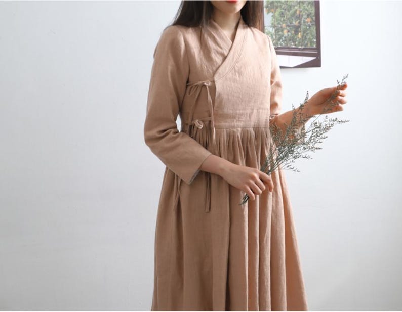 Puede incluir: Un vestido largo de lino de color marr&oacute;n claro con cuello en V y un lazo en la cintura. El vestido tiene una falda plisada y lo lleva una persona que sostiene un ramo de flores secas.