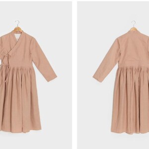 Puede incluir: Un vestido largo de color rosa claro con mangas largas, cuello de envoltura y falda fruncida.