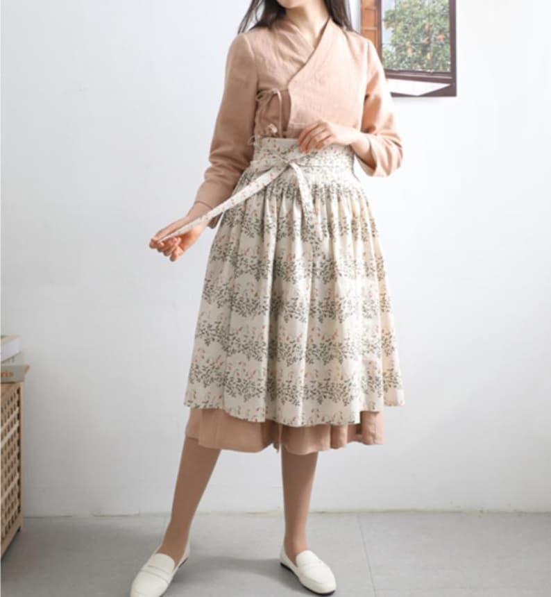 Puede incluir: Una mujer con un top marr&oacute;n claro de manga larga y una falda blanca con estampado floral. La falda tiene un cintur&oacute;n marr&oacute;n ancho con una larga corbata.