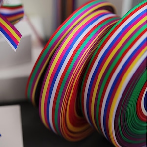 May include: Two rolls of multi-colored ribbon featuring a repeating pattern of vertical stripes in red, yellow, green, blue, and white. The ribbon has a smooth, satin-like texture and is coiled into tight, circular spools for storage and use.