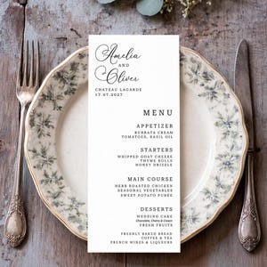 Peut inclure: Un menu de mariage avec les noms "Amelia et Oliver" en écriture cursive. Le menu détaille les entrées, les plats, les plats principaux et les desserts. La carte est posée sur une assiette à motifs floraux, avec une fourchette et un couteau.