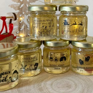 Peut inclure: Une collection de petits bocaux en verre remplis d'un liquide clair, chacun avec un couvercle en métal doré et des graphismes et textes de fêtes imprimés en noir, dont Meilleurs Voeux, Joyeux Noel 2023, Bonne Annee 2024 et Bonnes Fetes.