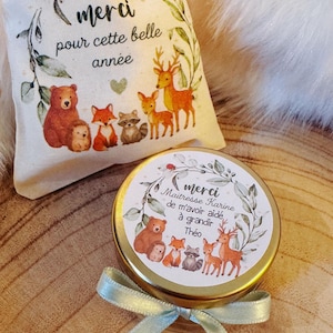 Peut inclure: Un petit pot rond avec un couvercle doré et un ruban bleu clair, à côté d'un petit sac en tissu. Les deux articles présentent des illustrations d'animaux de la forêt et le mot français « merci ». Le sac porte l'inscription « merci pour cette belle année ».