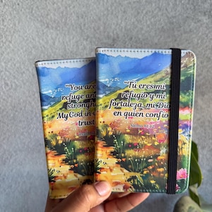 May include: Two phone cases with a landscape design featuring mountains, flowers, and text. The cases have a colorful design with the text "You are refuge and stronghold, My God in trust, Psalm 91" and "Tú eres mi refugio y mi fortaleza, mi Dio en quien confio, Salmo 91:12".