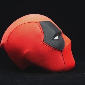 May include: A red and black Deadpool mask, a superhero costume accessory. The mask is primarily red with a black eye covering and white eye details. The mask is set against a black background.