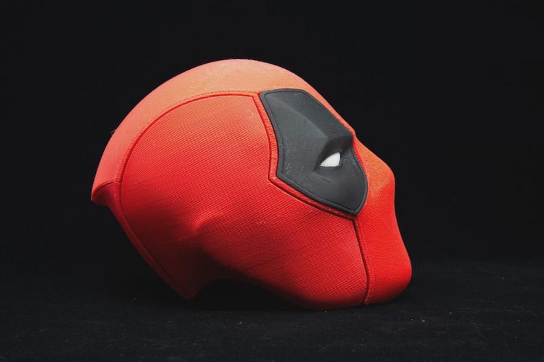 May include: A red and black Deadpool mask, a superhero costume accessory. The mask is primarily red with a black eye covering and white eye details. The mask is set against a black background.