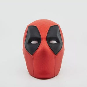 May include: A red Deadpool mask with black eye and mouth details. The mask has a textured surface and is set against a white background. The eye openings have a mesh material.