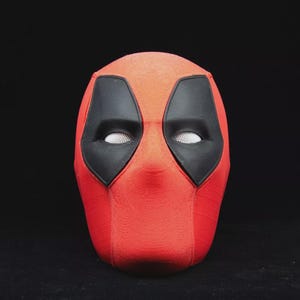 May include: A red Deadpool mask with black eye coverings and white mesh eye openings. The mask is a full head covering, with a textured surface. The mask is a costume accessory.