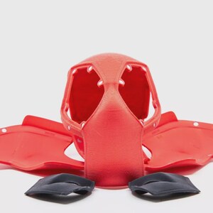 May include: A red plastic helmet with a face guard and two side pieces, along with two black pads. The helmet has multiple openings and white dots. The pieces are arranged on a white surface.