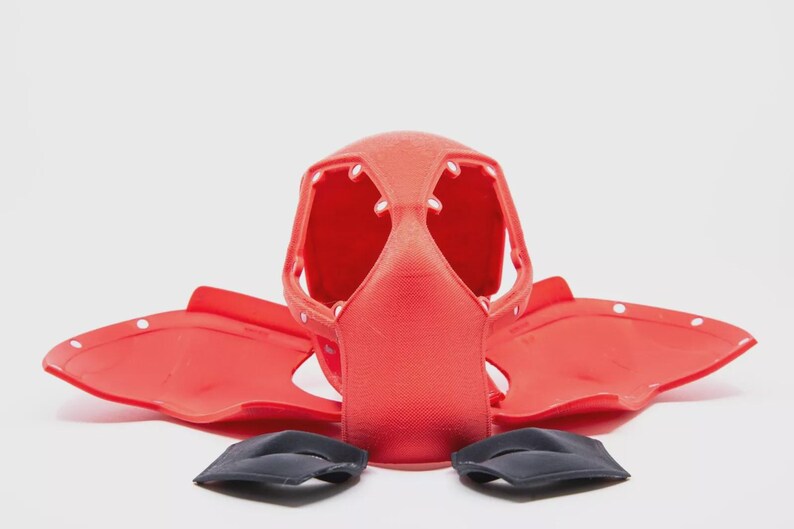 May include: A red plastic helmet with a face guard and two side pieces, along with two black pads. The helmet has multiple openings and white dots. The pieces are arranged on a white surface.