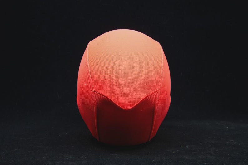 May include: A red, fabric-covered object with a rounded top and a pointed, V-shaped bottom. The object is set against a black background, with the color gradient shifting from a darker red at the base to a lighter shade at the top.