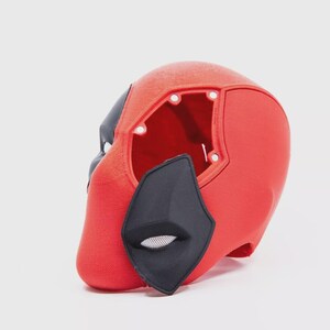 May include: A red and black Deadpool mask, a superhero costume accessory. The mask is primarily red with black accents around the eyes and mouth. The mask has an open back and is set against a white background.