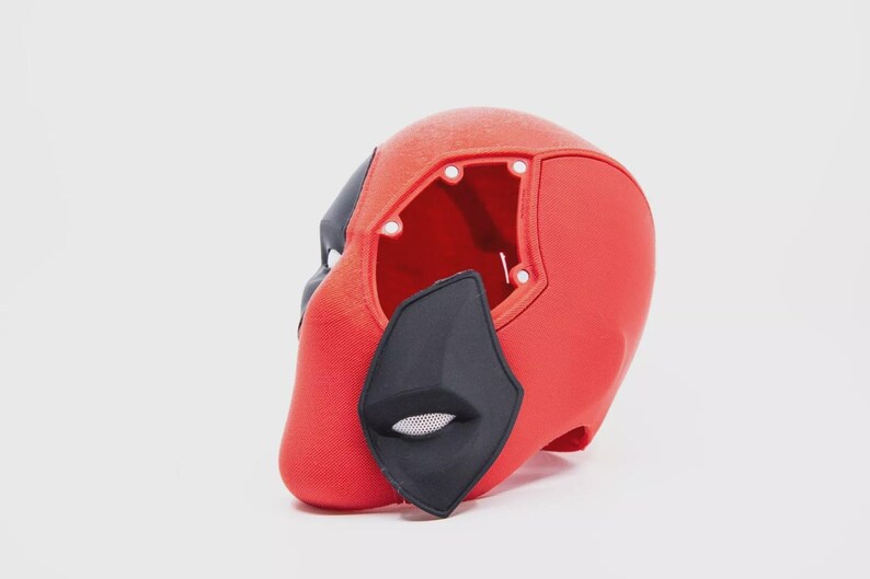 May include: A red and black Deadpool mask, a superhero costume accessory. The mask is primarily red with black accents around the eyes and mouth. The mask has an open back and is set against a white background.