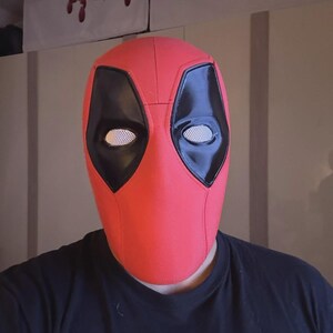 May include: A red Deadpool mask with black eye coverings and white mesh eye inserts. The mask is made of a textured material and is designed to cover the entire face. The mask is a cosplay item.