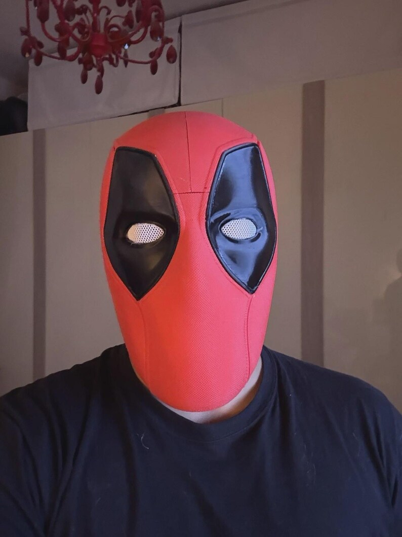 May include: A red Deadpool mask with black eye coverings and white mesh eye inserts. The mask is made of a textured material and is designed to cover the entire face. The mask is a cosplay item.