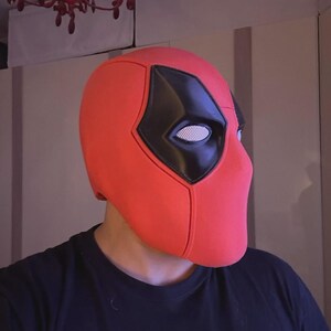 May include: A red Deadpool mask with black eye coverings and a white mesh eye area. The mask is a full head covering, and the design is based on the comic book character. The mask is made of a smooth material.