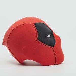 May include: A red Deadpool mask with black eye coverings and white eye details. The mask is a solid color and has a textured surface. The mask is a replica of the character's mask from the Marvel Comics.