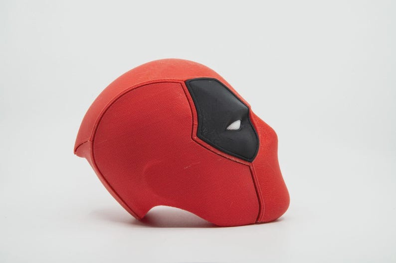 May include: A red Deadpool mask with black eye coverings and white eye details. The mask is a solid color and has a textured surface. The mask is a replica of the character's mask from the Marvel Comics.