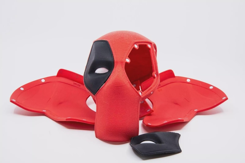 May include: A red and black Deadpool mask with matching red ear pieces and a separate black eye mask. The mask has a mesh eye covering and is made of a textured material. The ear pieces have white button-like details.