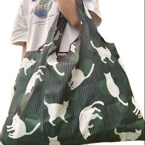 May include: A green and white patterned tote bag featuring white cat silhouettes on a background of green stripes and circles. The bag has brown trim and handles. The bag is being held by a person wearing a white t-shirt.