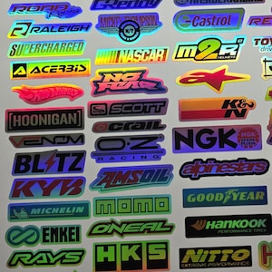 May include: A collection of holographic stickers featuring various brand logos and graphics. The stickers include logos for racing, automotive, and performance brands, with vibrant colors and reflective surfaces.