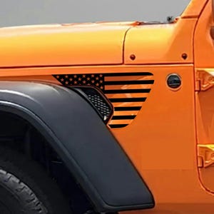 May include: Orange vehicle with a black and orange American flag graphic on the side panel. The flag design features black stripes and stars, adding a patriotic touch to the car's exterior. The vehicle's tire and fender are also visible.