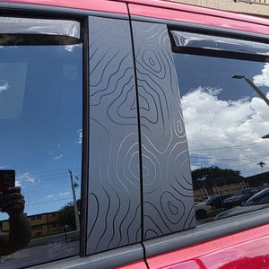May include: Red vehicle with black window trim. The window trim features a topographic map design in gray and white. The windows reflect the sky and surrounding environment.