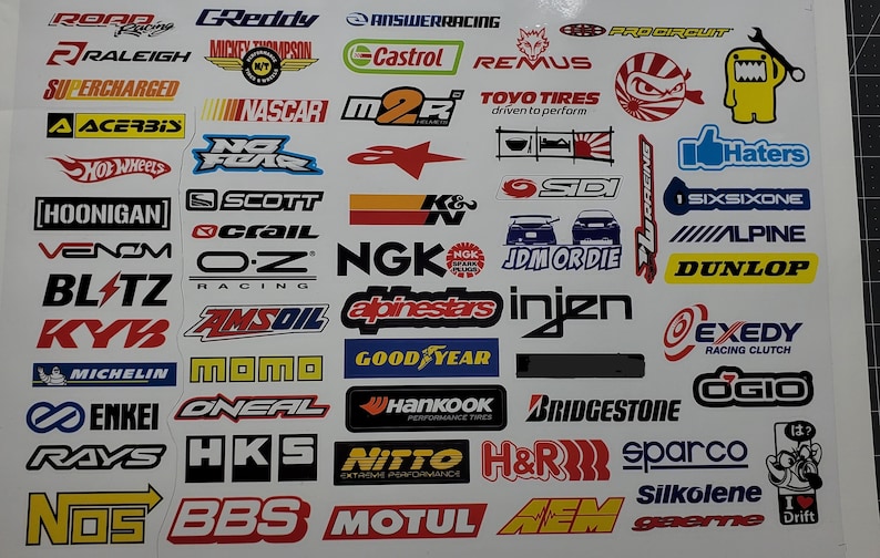 May include: A sheet of colorful stickers featuring various car and racing brands, including ROR, GReddy, Answer Racing, Castrol, Remus, NASCAR, K&N, NGK, AEM, HKS, and more.