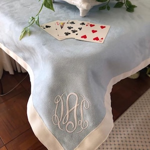 May include: A light blue table cover with a cream-colored border and embroidered monogram. Playing cards with red and black suits are on the table. A white planter with greenery is in the background.