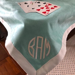 May include: A light teal table runner with a white border and a monogram. Playing cards with red hearts and black clubs are on the runner. A white decorative vase sits on the runner.