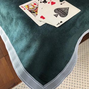 May include: A dark teal green cloth with a white and blue striped border. Playing cards are fanned out on the cloth, including the Queen of Diamonds, the Seven of Clubs, the Five of Hearts, and the Ace of Spades.
