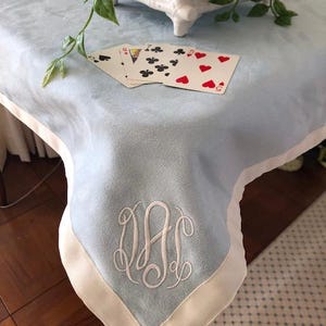 May include: A light blue tablecloth with a white border and a white monogram in the center. The monogram is the letters "DAC". There are playing cards on the tablecloth.