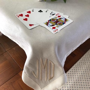 May include: A light-colored, monogrammed table cover with playing cards. The cards include the 8 of hearts, 2 of clubs, and the Queen of diamonds. The monogram features the letters JMD in a decorative font. The table cover has a satin border.