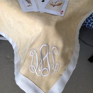 May include: A beige tablecloth with a white embroidered monogram and white ribbon trim. Playing cards are on the table. A blue and white decorative box is in the background.