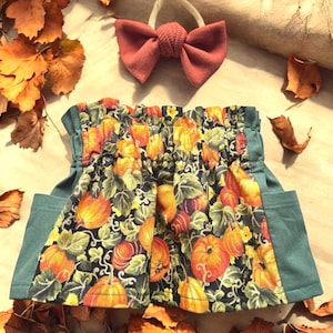 May include: A fall-themed skirt with a pumpkin print, featuring orange pumpkins and green leaves on a black background. The skirt has green side panels and is paired with a rust-colored bow.