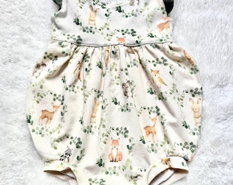 May include: A white baby onesie with a green and brown woodland animal print. The onesie has ruffle sleeves and a gathered skirt.
