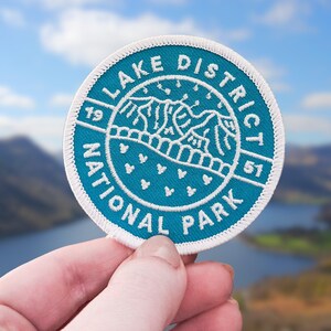 May include: A circular teal patch with white embroidery. It features the text "Lake District National Park" and a mountain range design. The years "19" and "51" are also included. The patch is held up against a blurred background of a lake and hills.