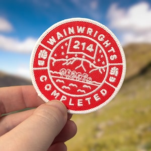 May include: A red and white embroidered patch with the text "Wainwrights Completed" and the number "214" in the centre. The patch features a stylised image of a mountain range with trees and a lake.
