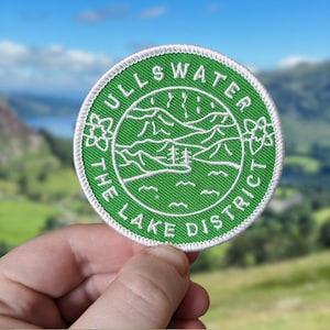 May include: A green embroidered patch with white stitching. The patch features a stylised image of a lake with mountains in the background. The text "ULLSWATER THE LAKE DISTRICT" is written in a circle around the image.