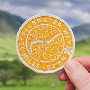 May include: Circular yellow patch with white embroidered text and a map design. The text reads "ULLSWATER WAY", "20 MILES 32 KM". The patch has a white border. The background is a blurred view of green hills and a blue sky.
